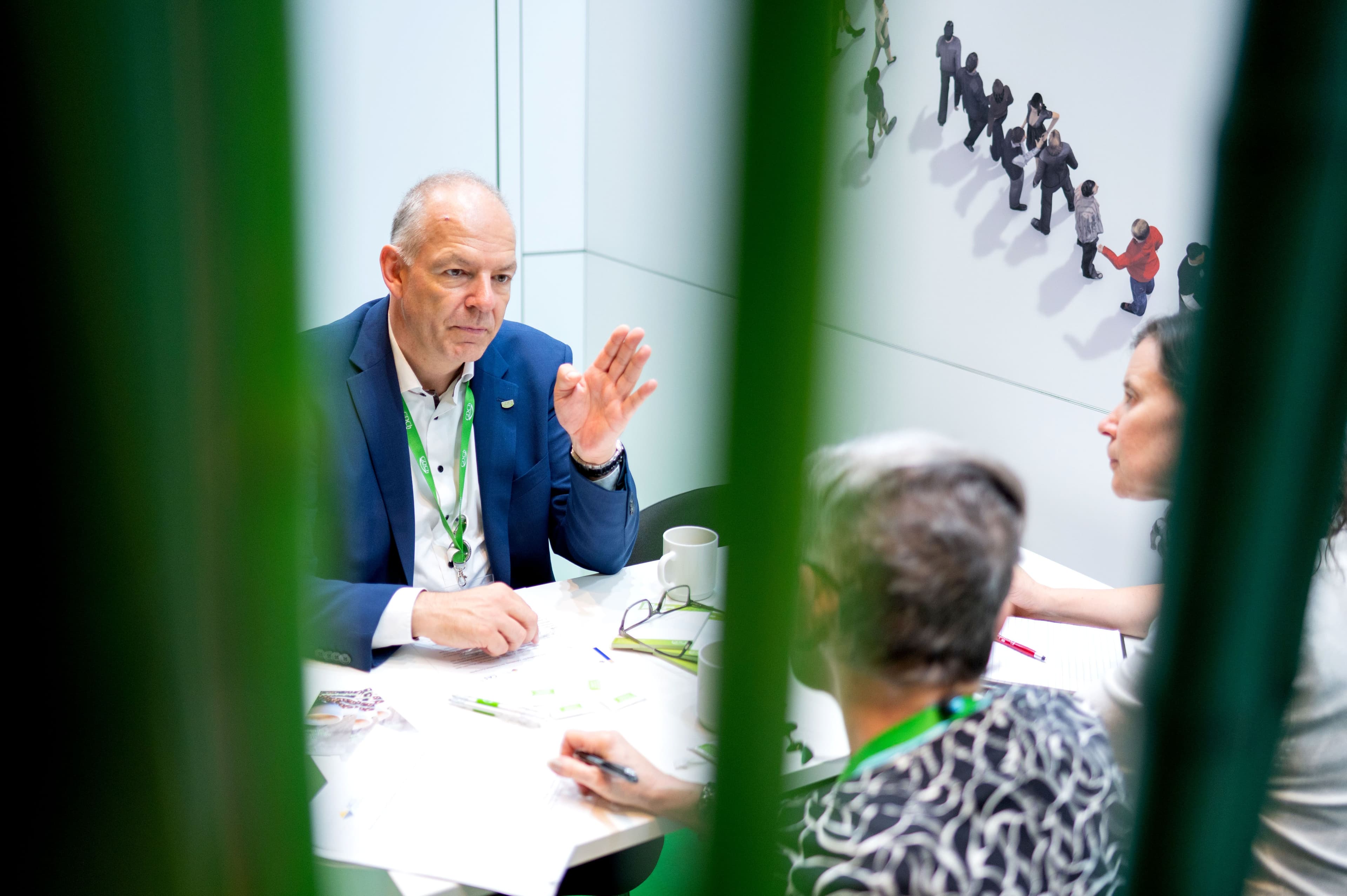 Blick durch grüne Pflanzen auf ein Gespräch in einem Besprechungsraum. Ein älterer Mann im blauen Anzug mit grünem Lanyard gestikuliert während er spricht. Er sitzt an einem weißen Tisch mit Kaffeetasse und Unterlagen. Ihm gegenüber sitzen zwei Personen mit dem Rücken zur Kamera. An der Wand hängt ein Poster mit Silhouetten von Menschen in einer Reihe. Die Aufnahme durch die Blätter im Vordergrund erzeugt einen natürlichen Rahmen.